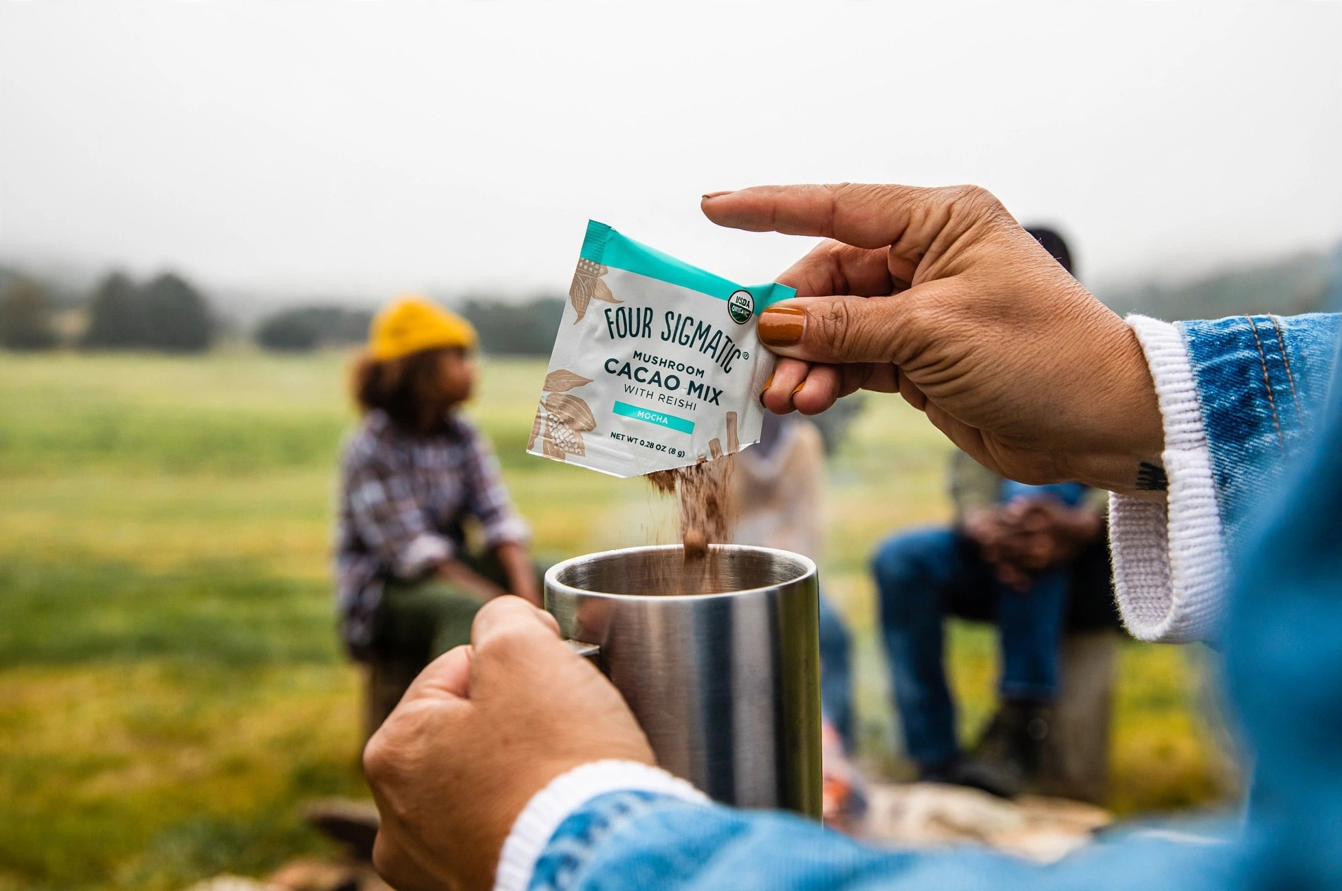 Hands pouring Four Sigmatic cacao mix into a metal mug outdoors.