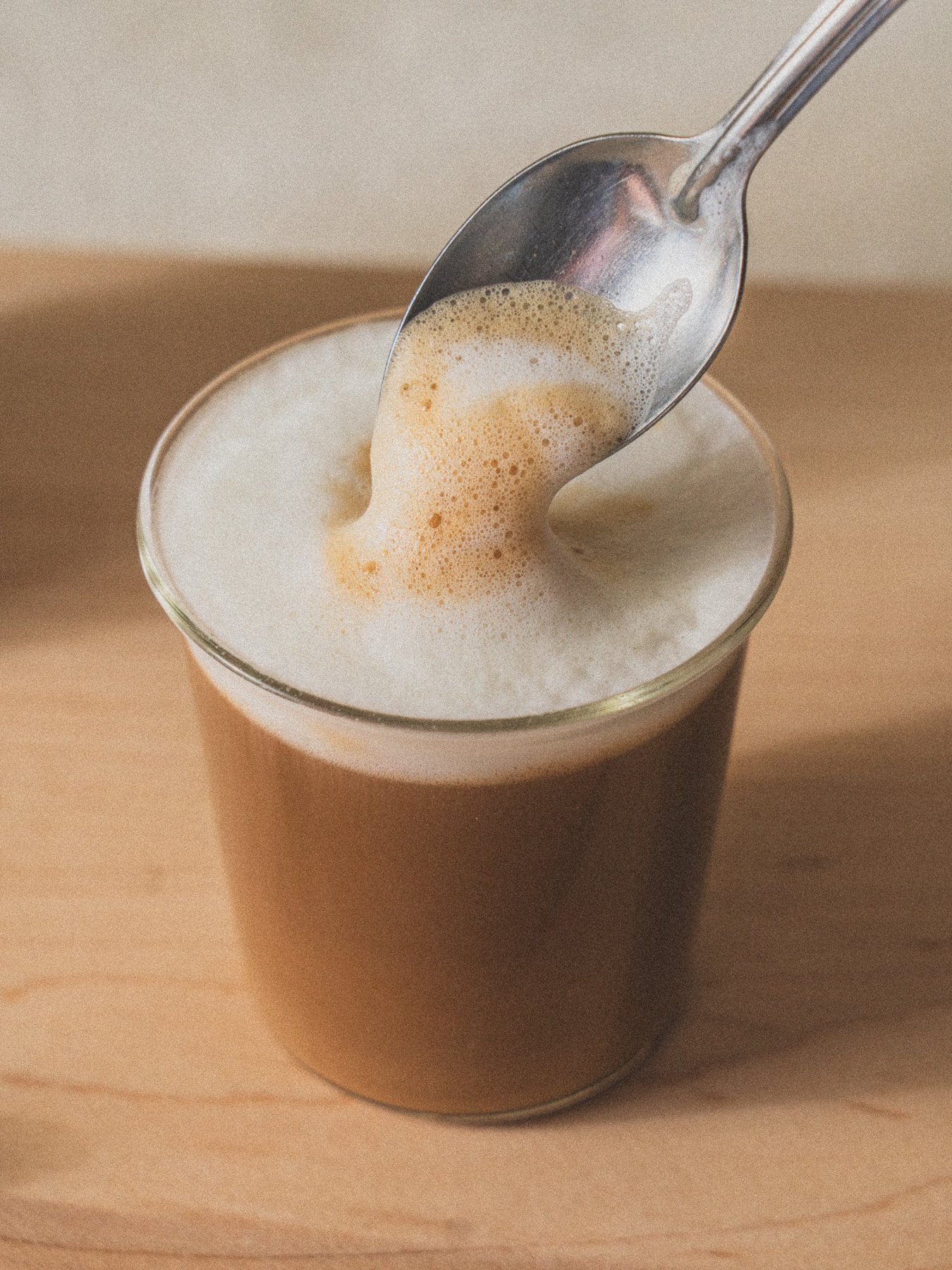 A spoon lifting frothy milk over a glass of latte on wood.